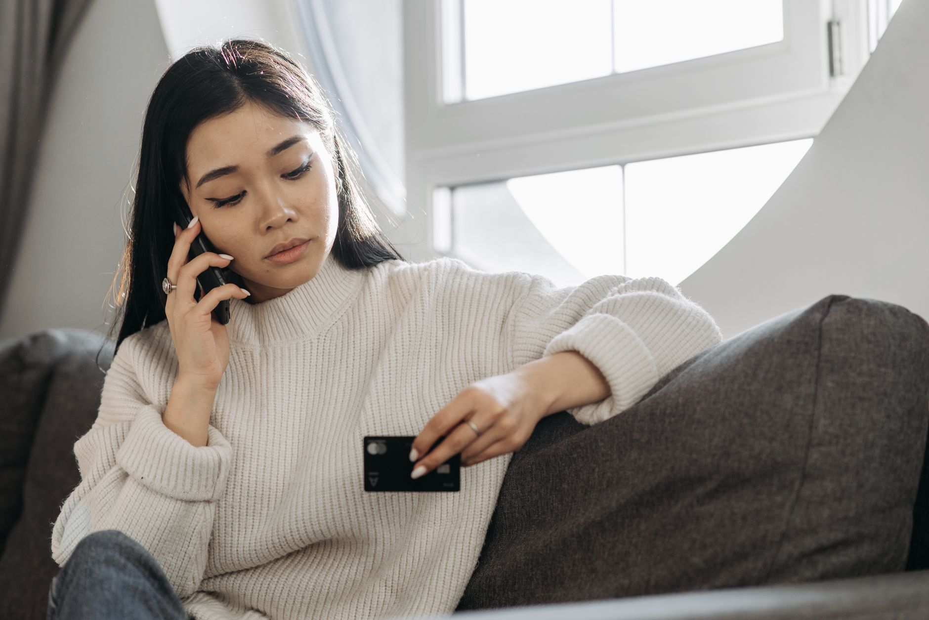 Une femme au téléphone, assise sur un canapé gris près d’une fenêtre, l’air concentré.