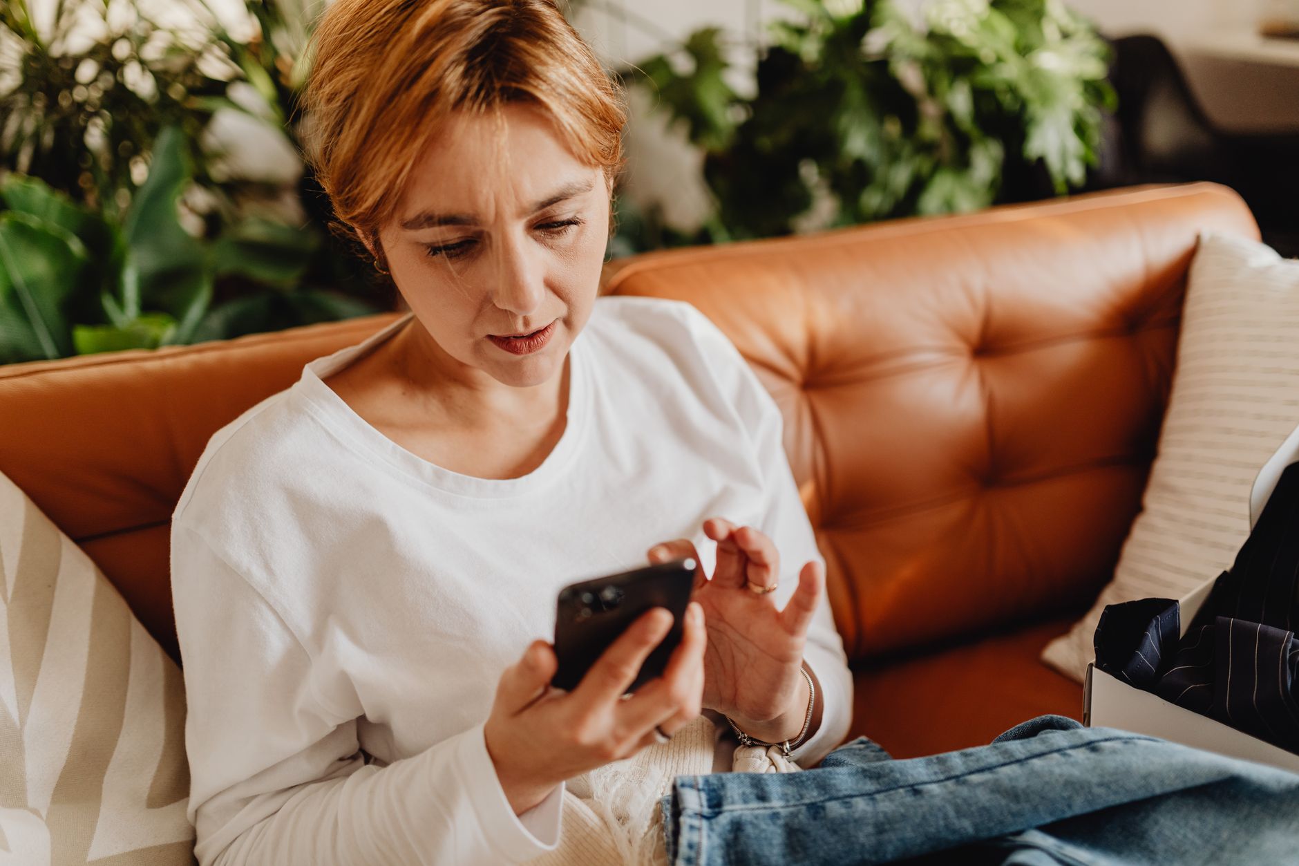 Une femme aux cheveux courts, assise sur un canapé en cuir, regarde attentivement l’écran de son téléphone.