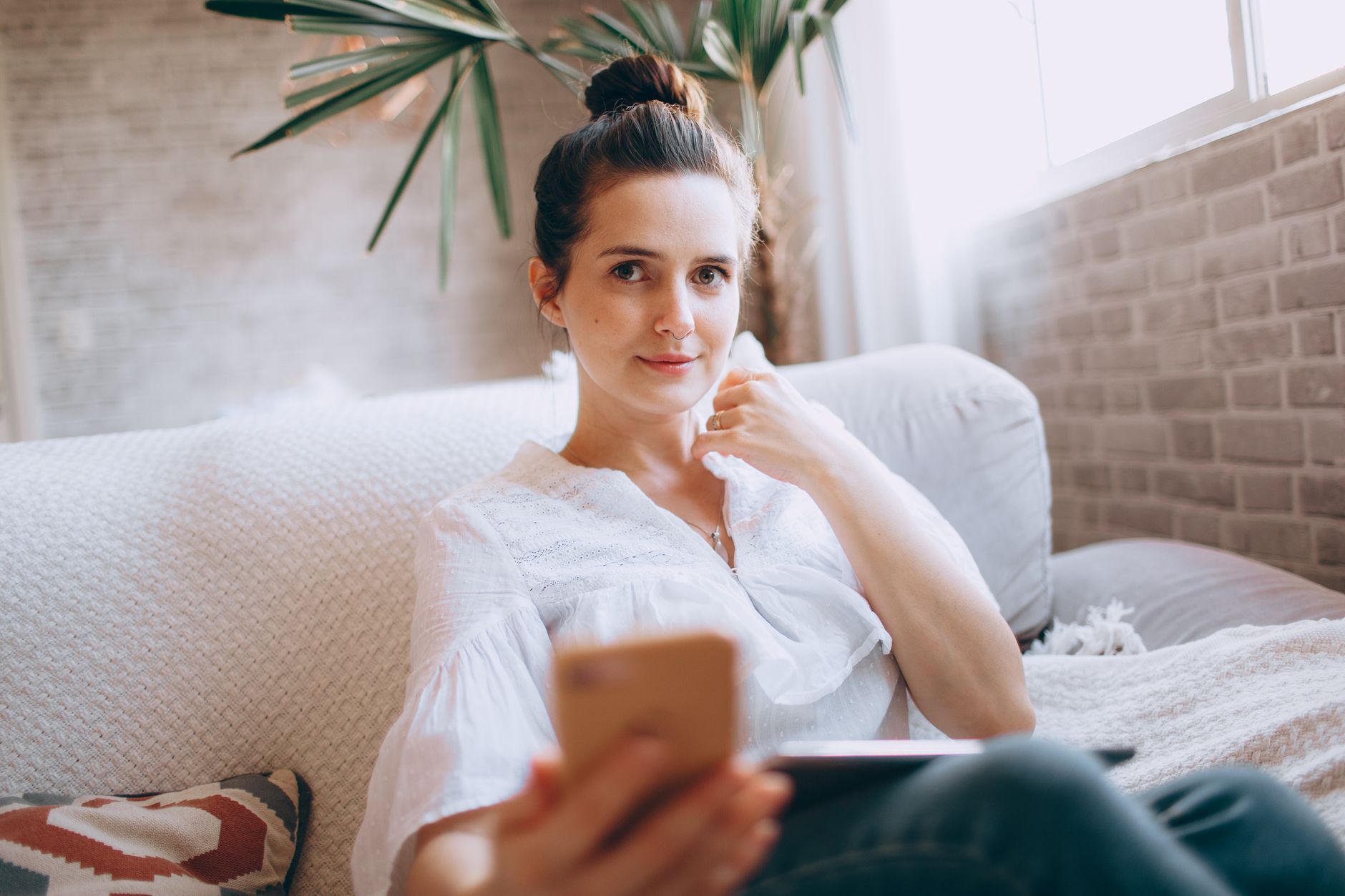 Une femme assise sur un canapé clair, un téléphone à la main, regarde vers l’objectif dans un salon lumineux.