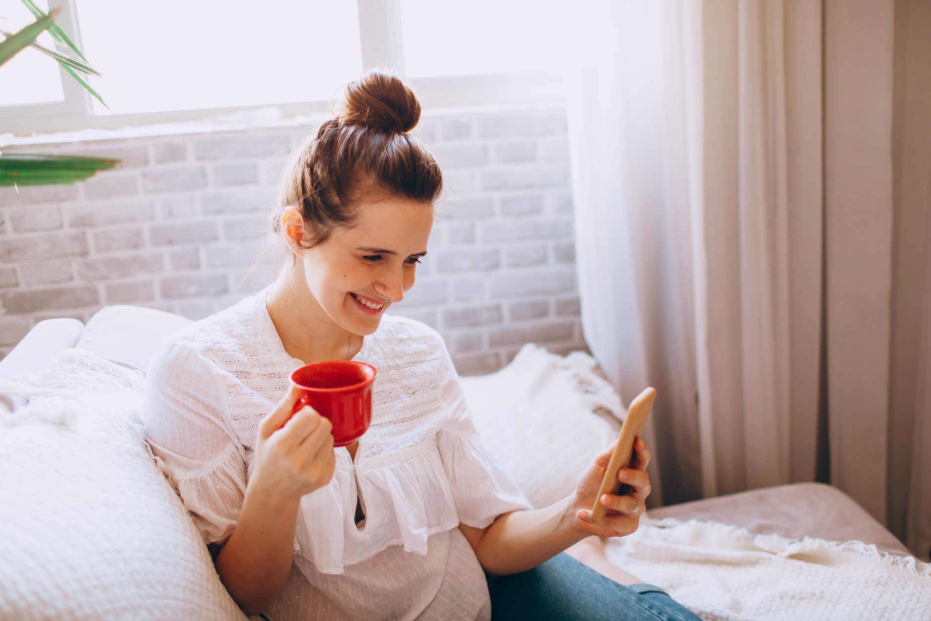 Une femme souriante, une tasse rouge dans une main et un téléphone dans l’autre, assise sur un canapé près d’une fenêtre.