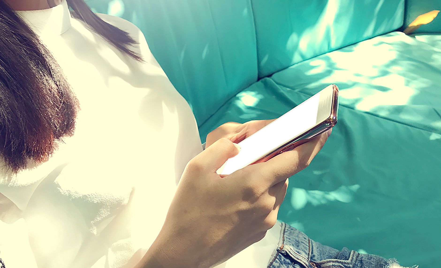 Gros plan sur une main qui tient un smartphone, sur une banquette turquoise éclairée par le soleil.