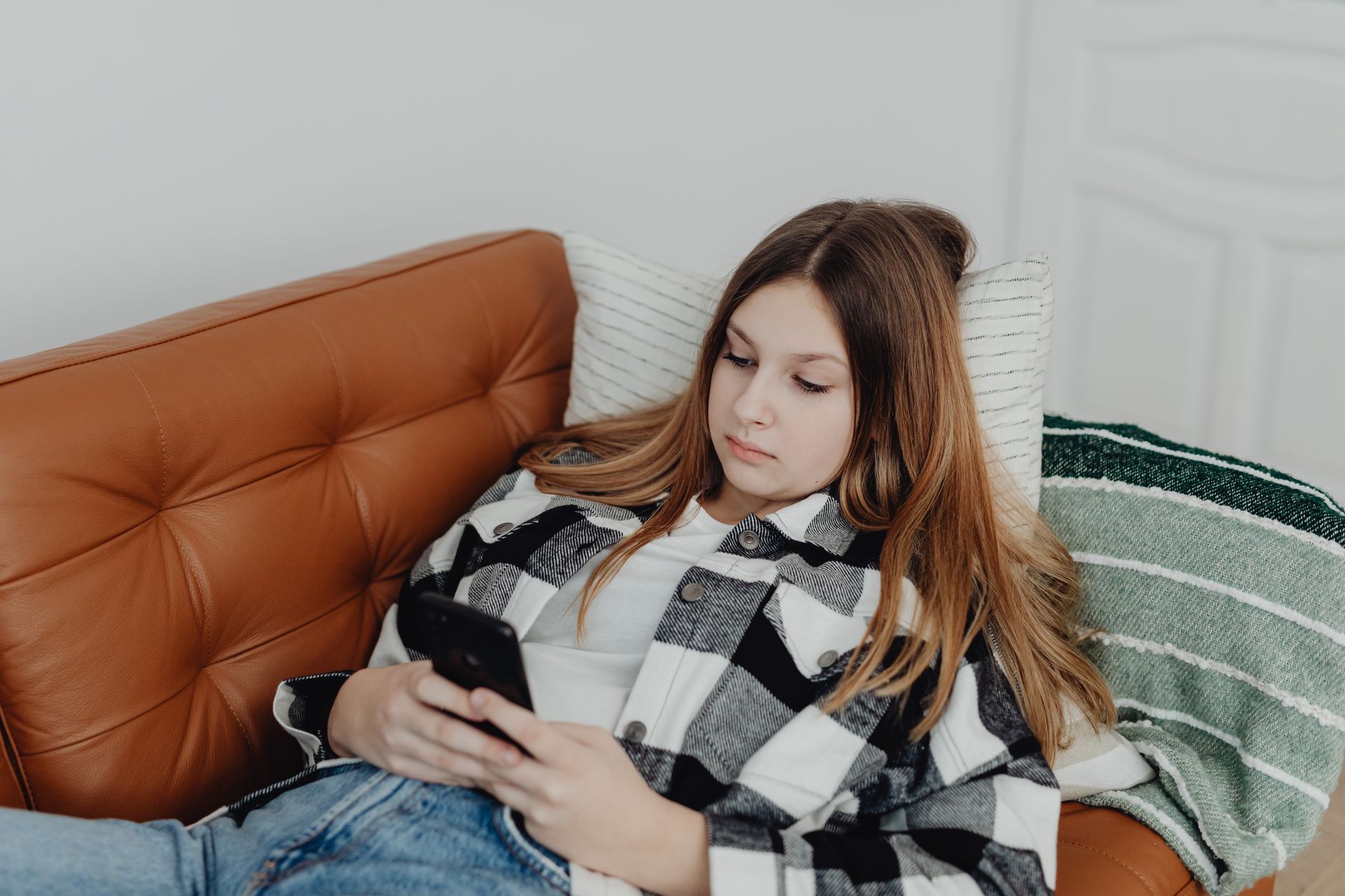 Une personne allongée sur un canapé en cuir, un téléphone entre les mains, dans un salon lumineux.